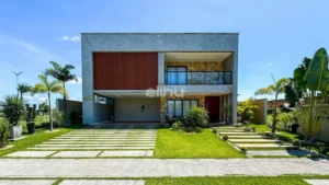 Imagem de frete de uma casa em estilo moderno com fachada quadrangular. Na imagem é possível visualizar dois andares, embaixo com uma entrada de garagem e janelas de sala e no andar superior, uma varanda do que seria a suíte principal da casa